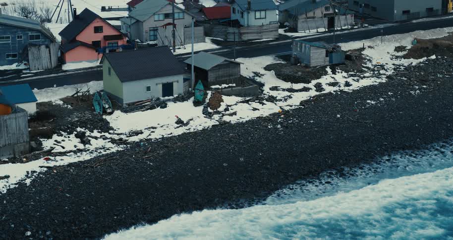 An overhead view captures a snow dusted rocky coastline with waves crashing onto the shore featuring colorful houses and small boats near a road