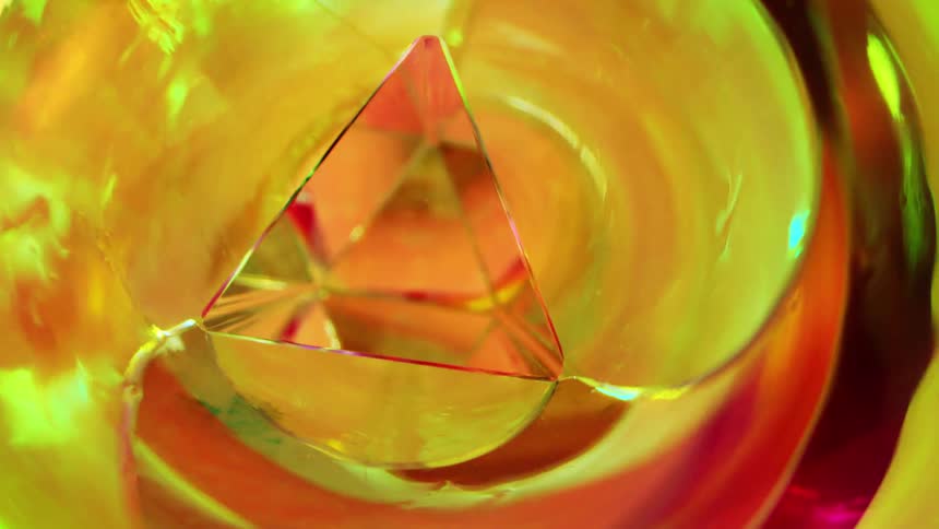 A pullfocus shot reveals an extreme closeup view into a kaleidoscope showing a central triangular prism reflecting swirling warm colors