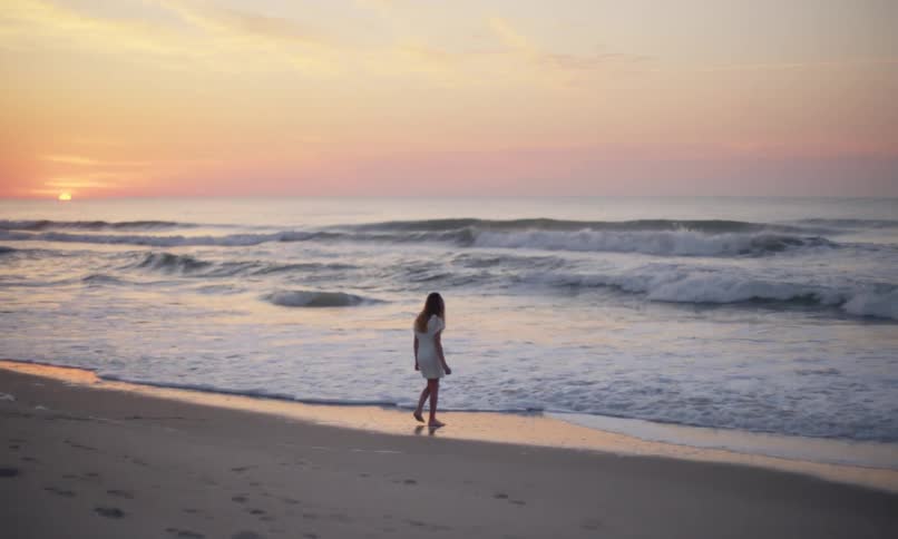 A young girl walks alone along the shoreline of a beach with a seagull scuttling nearby as the waves roll in and the sun kisses the horizon to create a serene rainbow sky at dusk.