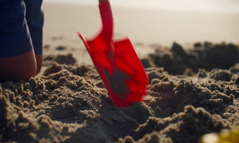 Close up of children digging holes in the sand with plastic shovels at a beach on a sunny day.