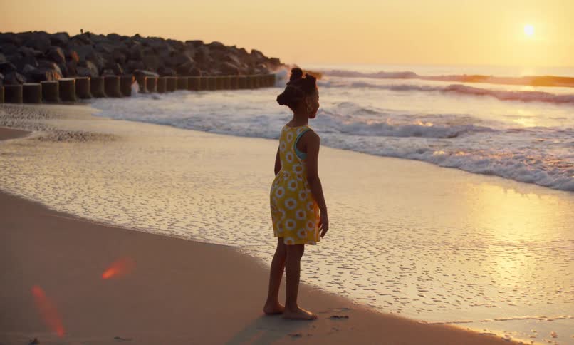 A little black girl in a yellow-flowered dress stands quietly at the edge of the water on a beach looking out at the golden sunset over the ocean waves and bends down to gently touch the incoming foamy surf with her hand.