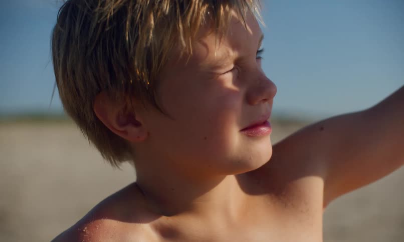 A young, shirtless, blonde boy holds his arm up to shade his eyes as he squints at the sun on a sandy beach.