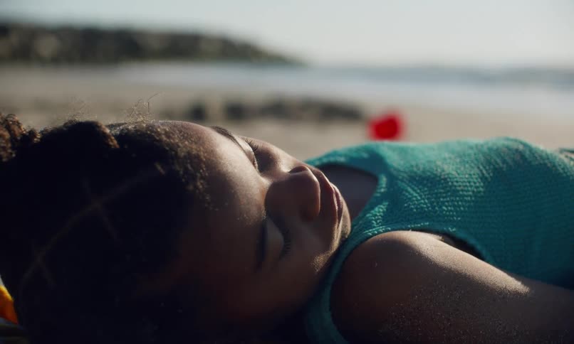 Close up of a young black girl laying on the sand and soaking up the sun at the beach as two other kids run and do cartwheels in the background.