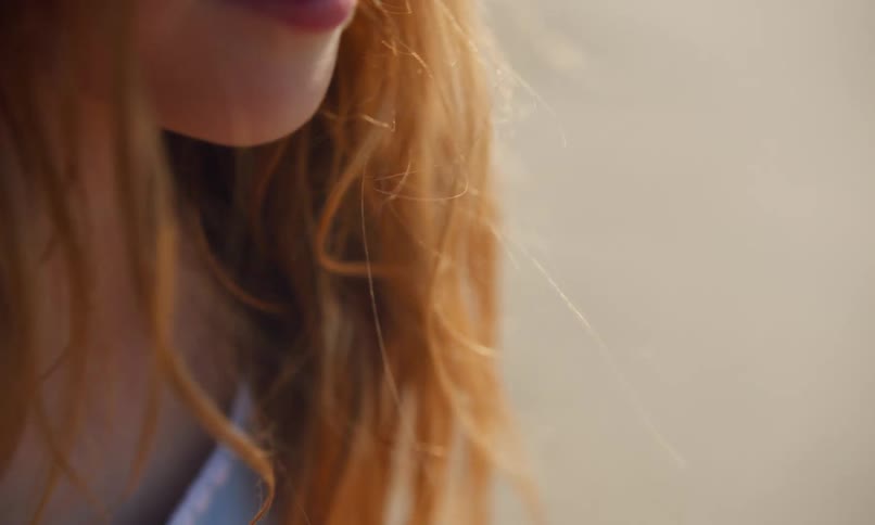 Close up of the lower face and shoulders of a young girl with the breeze blowing her red hair as the evening sun gives it a copper glow.