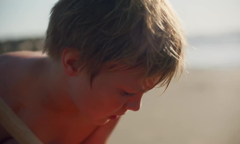 A little shirtless blonde boy is squatting down playing in the sand at the beach on a sunny day next to a little black girl in a blue swimsuit.