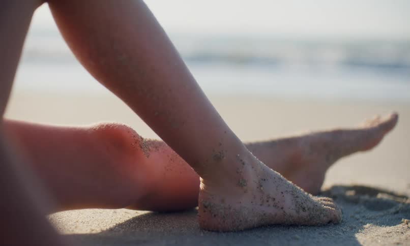 Lower legs and feet of a person laying out suntanning on the sand at the beach as the gentle surf comes in near them.
