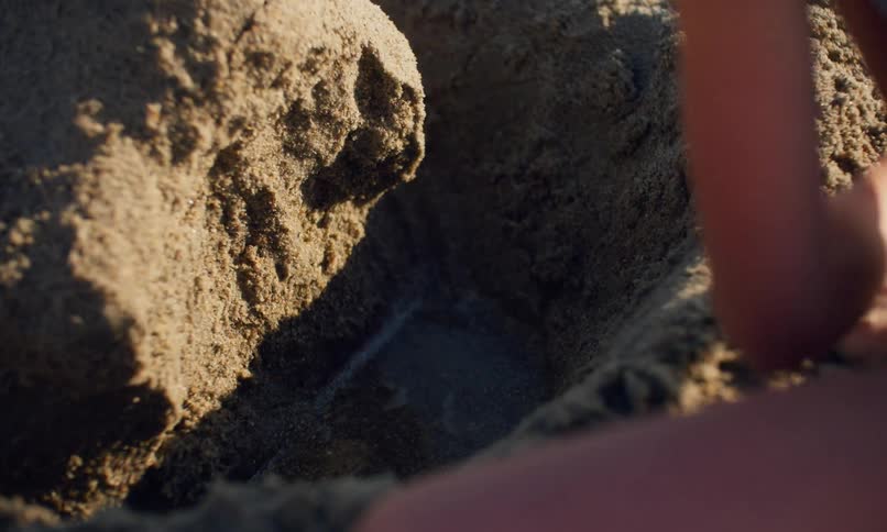 A young girl with long blonde hair is at the beach on a sunny day digging a hole in the sand with water inside and creating a mini sand hill and river canyon to play with.