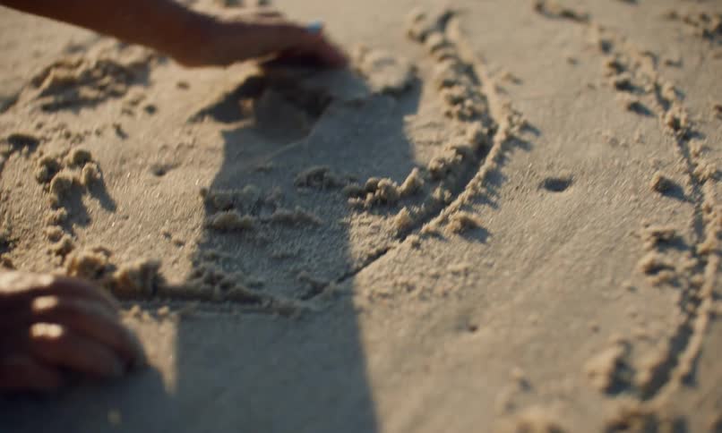 A little girl of color kneels in the sand at the beach on a sunny evening and uses her hands and fingers to draw shapes in the sand and then smooth and pat it down.