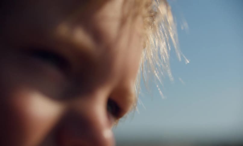 A blurry closeup captures the upper portion of a young blonde boy's face as the sea breeze gently blows his hair and he squints into the sun on a clear summer day.
