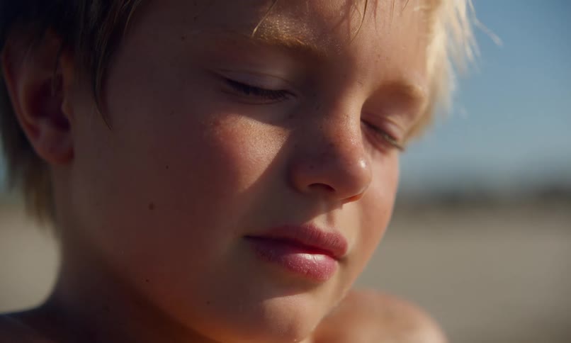 Closeup of a young, shirtless, blonde boy with his eyes closed basking in the warm sunlight and gentle sea breeze on a sandy beach.