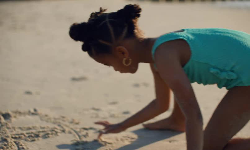 A young black girl child is happily moving around on her hands and knees at the beach as she draws letters and shapes in the sand with her finger.