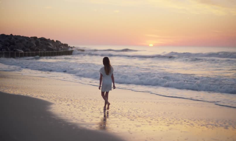 A lone, young girl in a white dress slowly walks along the shoreline of a beach gently kicking the surf as it touches her feet and watching the sun set on the horizon.