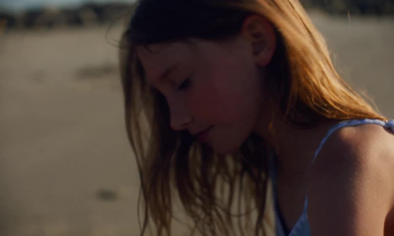 Close up of a young, red-headed girl in a blue swimsuit who is digging in the sand at the beach with a red plastic shovel at sunset.