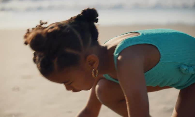 A young black girl in a blue swimsuit at the beach is kneeling and drawing shapes in the sand with a shell as the waves and surf roll in behind her.