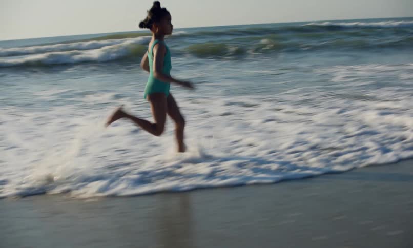 A little black girl wearing a teal swimsuit runs in the surf in the evening sun as the waves come in along the beach.