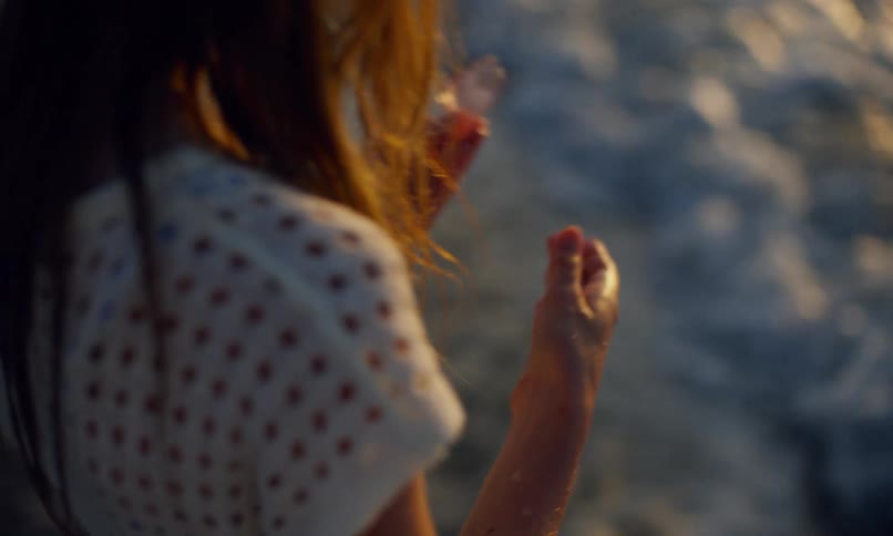 Close up of a young girl standing in the wet sand on the shoreline of a beach holding up a translucent piece of pink sea glass to the light of the setting sun to examine it and then bending over to wash it in the surf.