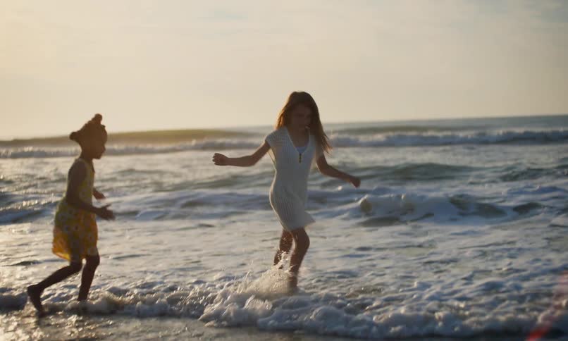 Slow motion of two diverse young girls with happy smiles running and chasing each other through the surf on a beach as waves roll in at sunset.