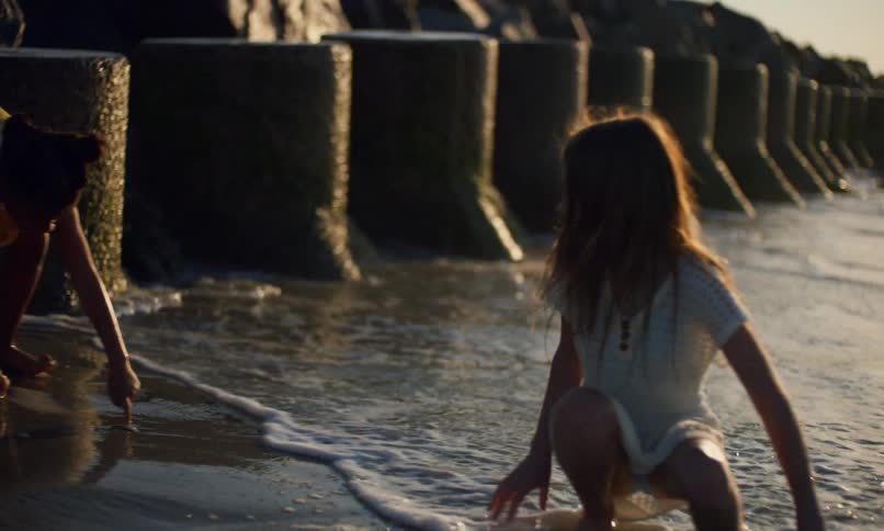 Slow motion of two young girls in summer dresses playing in the sand and surf along a rocky shoreline at sunset as ocean waves crash into stone barriers in the background.