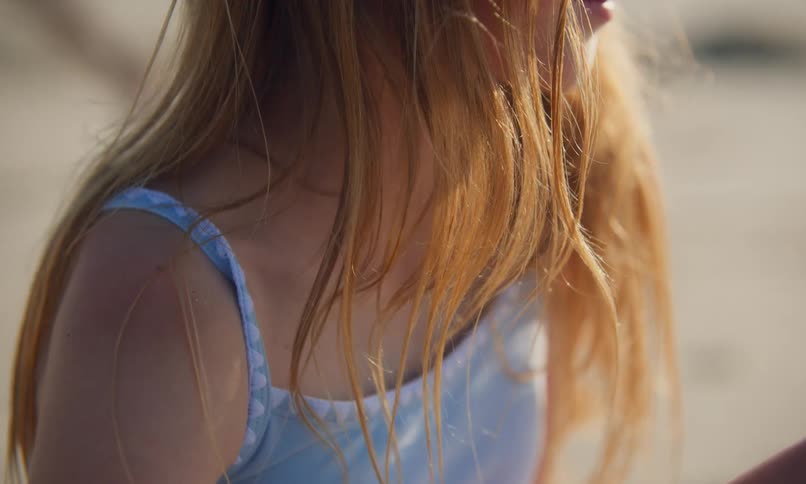 Close up of the lower face and shoulders of a girl with blonde hair sitting in the sun on a sandy beach.