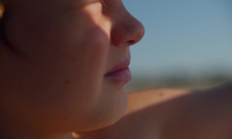 A young blonde boy sits in the sand on a beach and squints in the sun as he puts his arm up to shade his eyes.