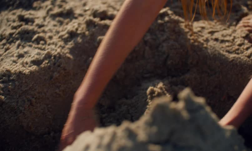 A young long-haired girl in a blue swimsuit is digging in the sand with her hands at the beach as the surf hits the shore behind her.