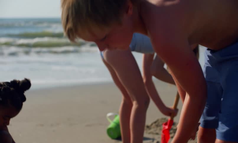 3 young kids, a boy and two girls, use plastic shovels and pails to dig holes and build sand castles at the beach on a sunny summer day.