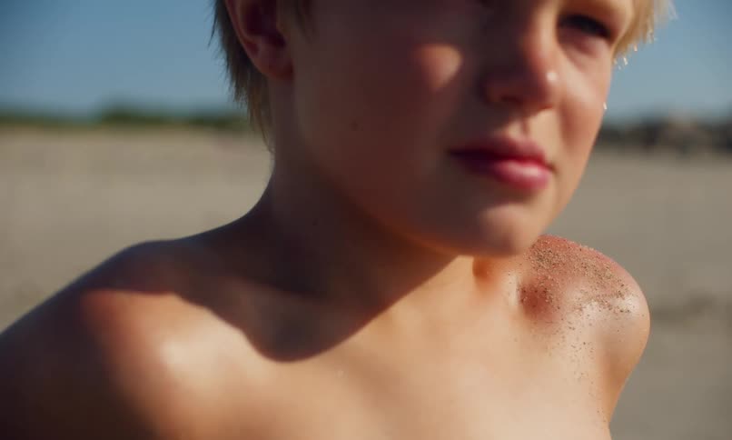 A young, shirtless, blonde boy sitting on the sand at the beach and soaking up the sun as he relaxes and looks around.