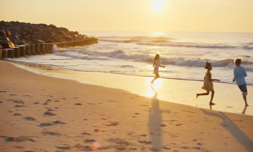 3 diverse children running and playing chase with each other along the shoreline of a sandy beach as gentle waves roll in at the height of sunset's golden hour.