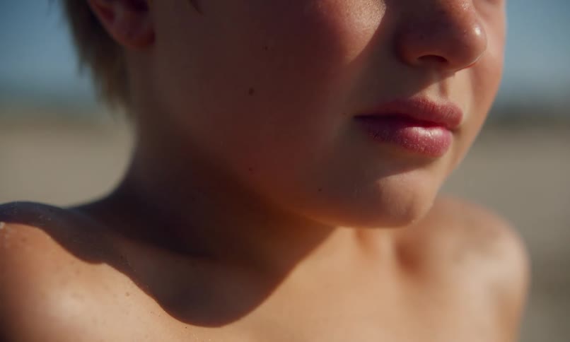 A young, shirtless, blonde boy sits in the sand enjoying the sun on a quiet beach and looks off into the distance and then to the side.