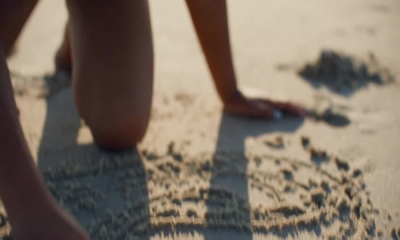 A young black girl child draws lines and shapes in the sand at the beach on a bright, sunny day.
