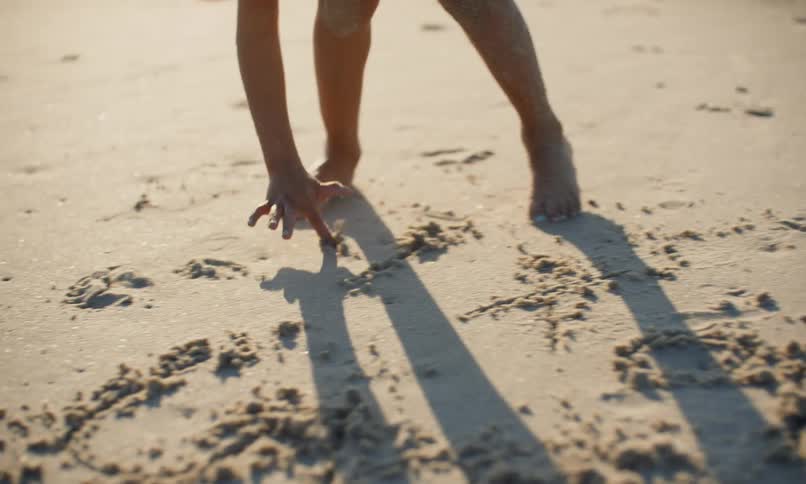 A young black girl with sandy hands and feet draws letters in the sand at the beach as the surf comes in behind her.