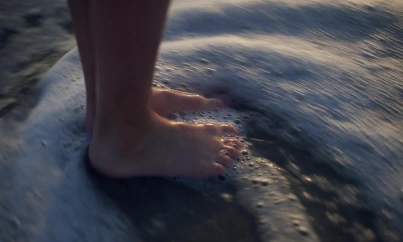 Close up view of a child's feet standing in the wet sand of a beach shoreline as the waves push the foamy surf in and the water swirls around their ankles then back out to sea.