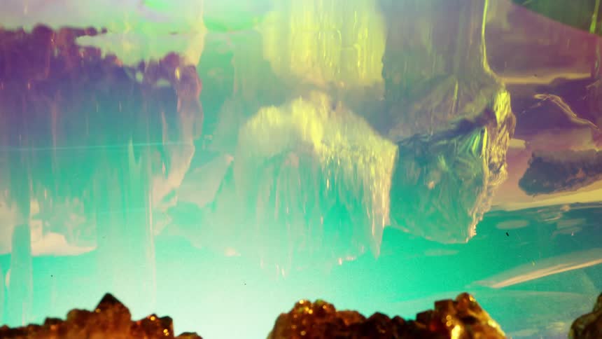 Vividly illuminated by blue and purple light several large roughhewn rocks are submerged with hints of yellow and orange light visible on the rippled reflections