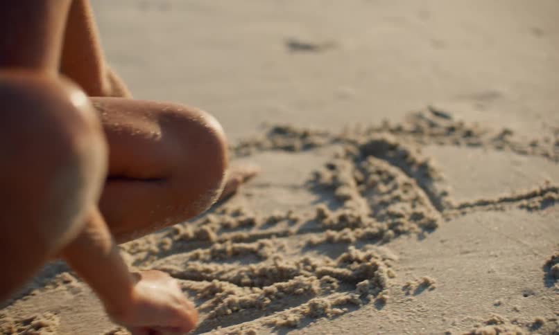 A little black girl in a blue swimsuit is on her hands and knees on the beach in the evening drawing a star shape in the sand with her fingers.