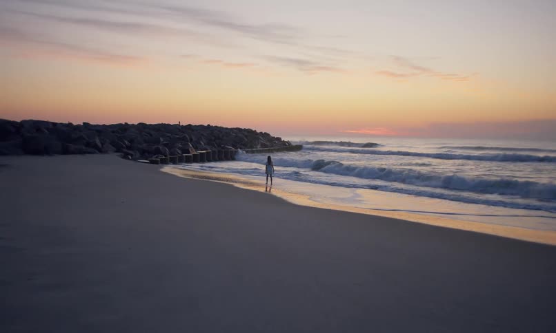 View of a female figure walking in the surf along the shoreline of a beach under a rainbow sky at sunset.
