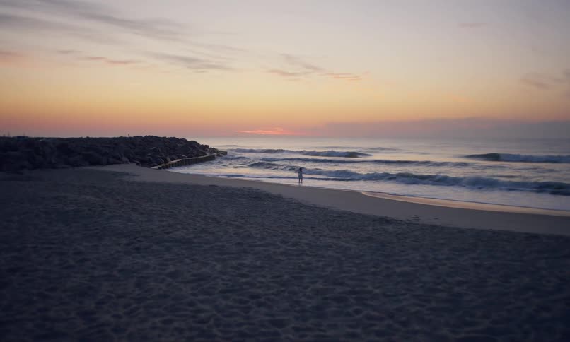 View from afar slowly moving toward a lone young girl walking along the shoreline of a sandy beach under a muted rainbow sky at dusk.