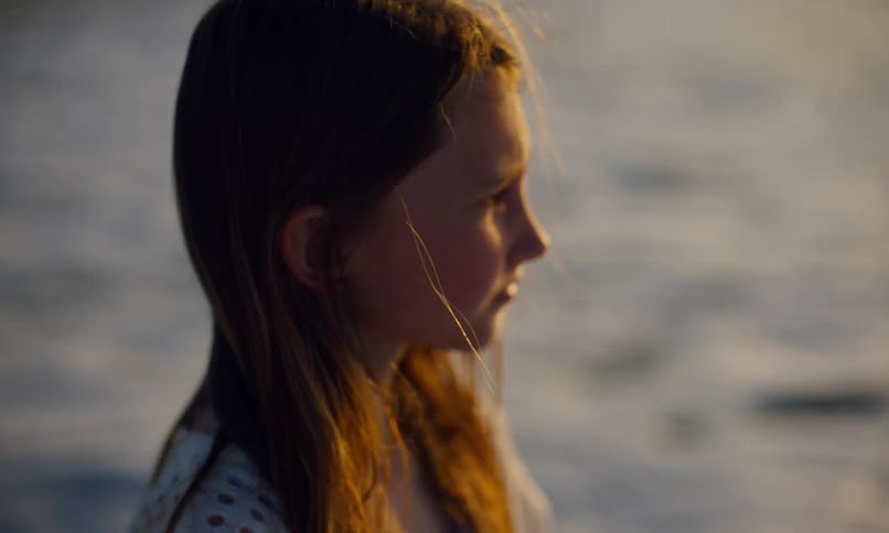 Close up of a young girl with long red hair and white dress standing on a beach who looks down at the surf while deep in thought and then up at the sunset on the horizon as the waves come in to the shore.