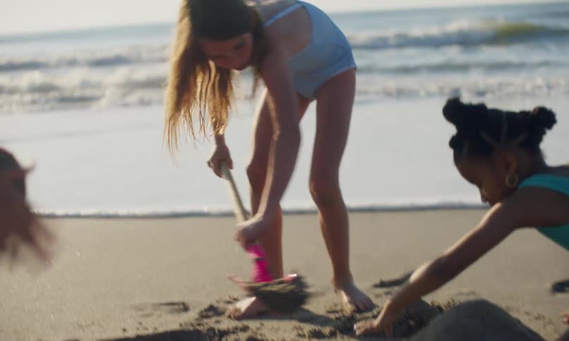 Two diverse young girls dig and play in the sand at a beach on a sunny day as waves push in the surf behind them.