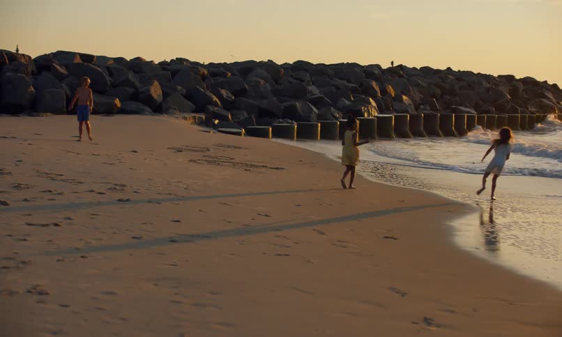 Three children at the beach running, playing chase, and splashing and falling in the surf as the waves bring the tide in and the sunset casts a golden glow over the ocean.