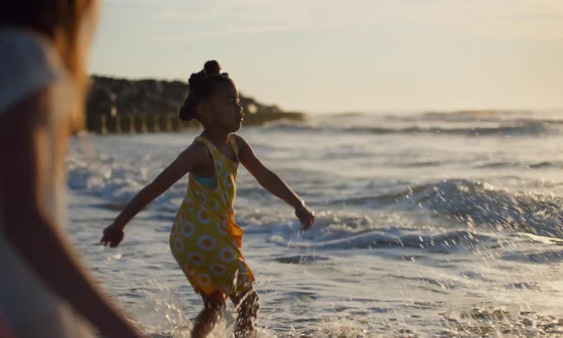 Slow motion of a young, black girl in a yellow dress playfully kicking the water as the waves roll in at sunset on the shoreline of an ocean beach, as another girl joins in next to her.
