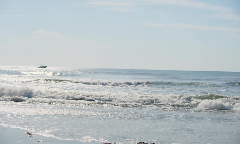 View of ocean waves crashing toward shore on a sunny beach in the early evening as a speedboat goes by in the distance and two female children walk by in the foreground carrying plastic shovel and pail.