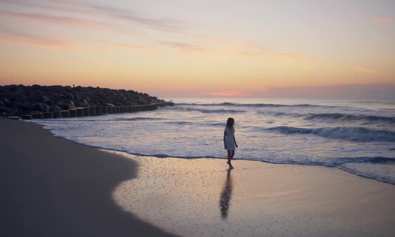 Drone footage zooming in and then out on a young girl on the beach at sunset enjoying the waves bringing the tide in as she walks alone on the shoreline.