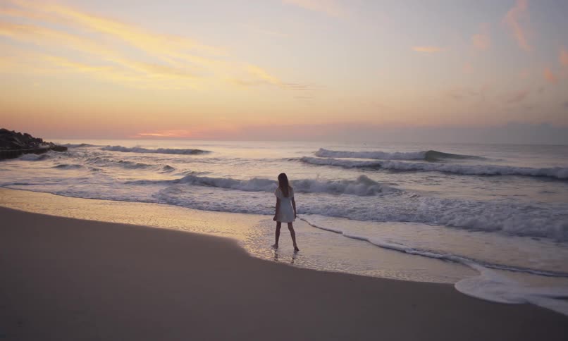 A young girl walking on the beach under a rainbow sky at sunset picks something up from the sand and throws it in the ocean as the waves and surf come in around her bare feet.