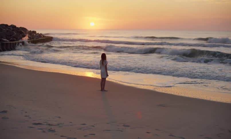 A young girl in a white dress is standing at the edge of the water on a sandy beach and quietly reflecting as she watches the sun set over the ocean waves.