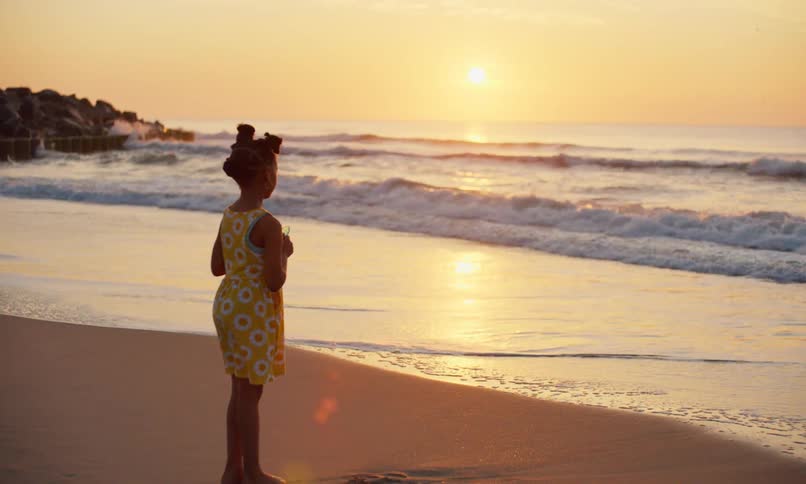A little black girl in a yellow-flowered summer dress stands alone in the sand of a beach at sunset peacefully watching the waves roll foamy surf on to the shore.