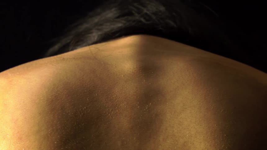 A closeup shot from behind shows the broad shoulders and upper back of a person with dark hair dimly lit against a black background