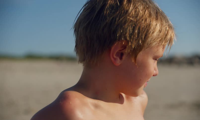 A shirtless, young, blonde boy sits in the sand on a sunlit beach gazing calmly into the distance and then turns to the side squinting into the sun and raises his arm to shade his eyes.