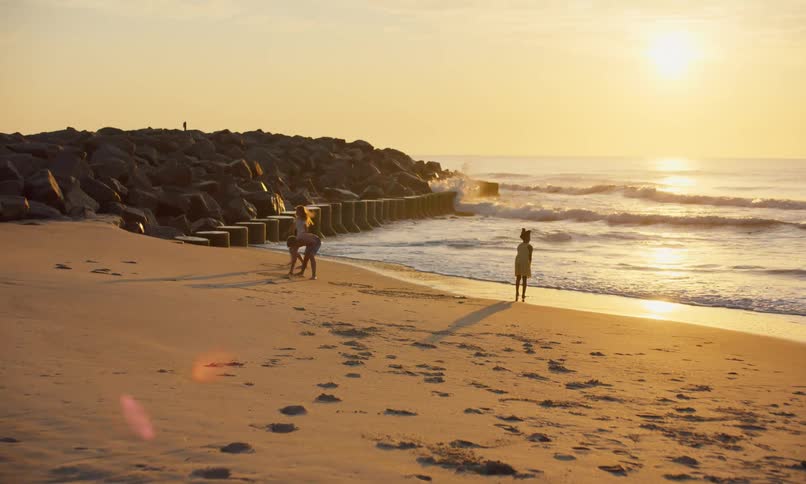 A beautiful golden sun sets on a sandy beach with waves crashing against a rocky jetty then rolling gently to the shoreline where three young kids are playing and exploring in the sand.