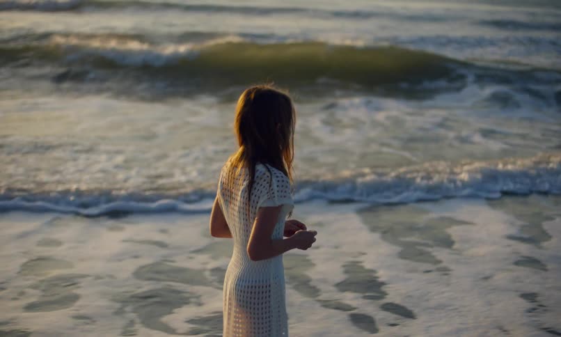 A young, long-haired girl in a white dress peacefully walking alone with bare feet in the surf as the waves roll in on a beach at sunset's golden hour.