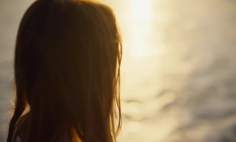 A young girl with long hair wearing a white swimsuit coverup dress is walking on the beach at sunset and then bends to pick up a piece of translucent sea glass from the wet sand, washes it off in the surf, and holds it up to the setting sun to examine it.